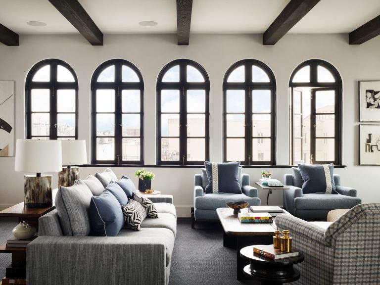A modern living room with five large arched windows, gray sofas decorated with blue and white pillows, two blue armchairs, a checkered chair, and a dark coffee table. The room features dark ceiling beams and neutral-toned walls with contemporary decor.
