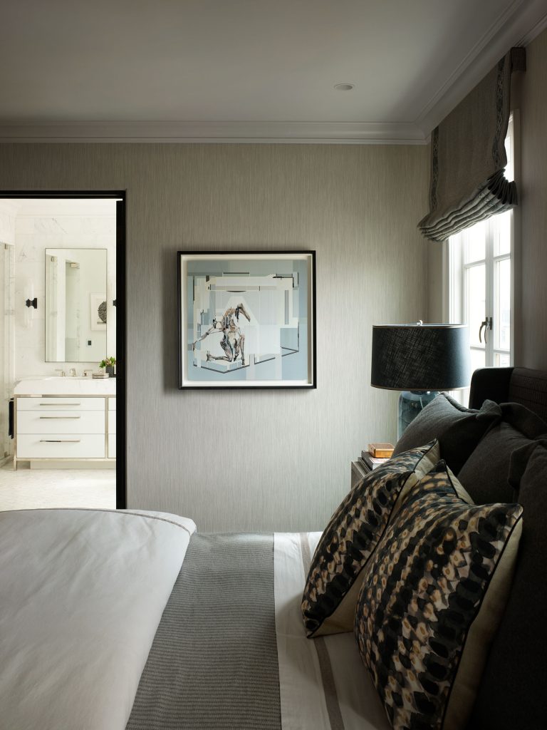 A modern bedroom with a neatly made bed, patterned pillows, and a dark lamp on a bedside table. A framed abstract artwork hangs on the wall. A window with a Roman shade lets in natural light. An open door reveals a bright, marble-tiled bathroom with a vanity.