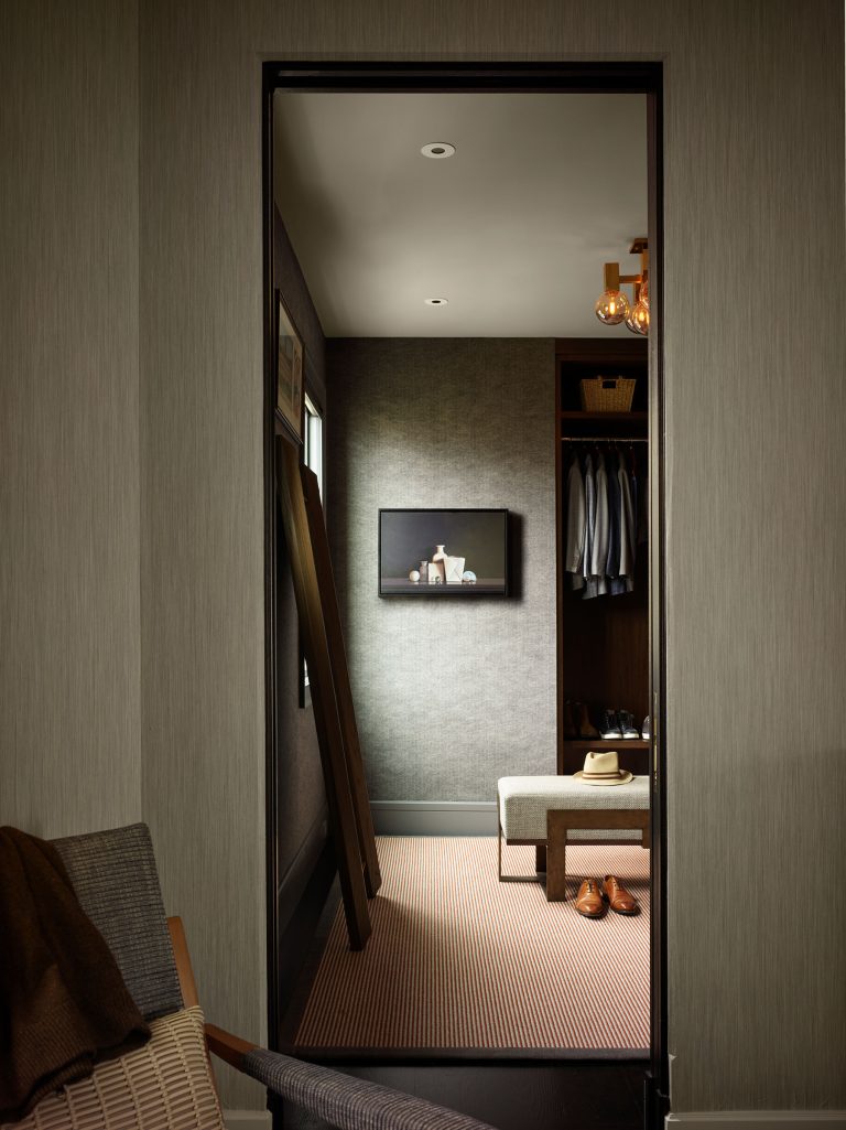 A walk-in closet seen through a doorway features a bench, a hat, brown shoes, hanging clothes, shelves with baskets, a painting on the wall, and soft lighting. A wooden ladder leans against the wall, and part of an upholstered chair is visible in the foreground.