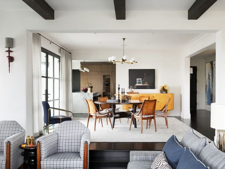 A modern open-plan living and dining room with exposed ceiling beams, plaid armchairs, a gray sofa, a wood dining table with six chairs, a large area rug, a brass light fixture, and wall art. Light pours in from tall French doors on the left.