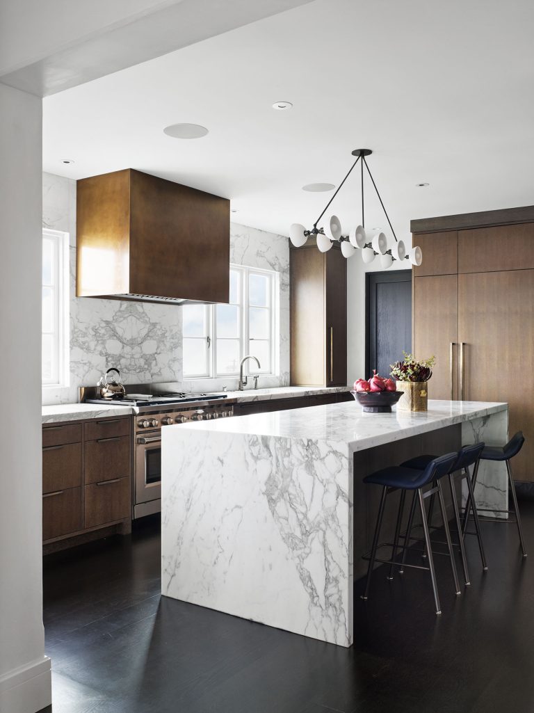Modern kitchen with a marble island and matching backsplash, wooden cabinets, dark floor, bronze range hood, and built-in appliances. Three black barstools are at the island, and a bowl of red apples sits on the counter. Contemporary chandelier overhead.