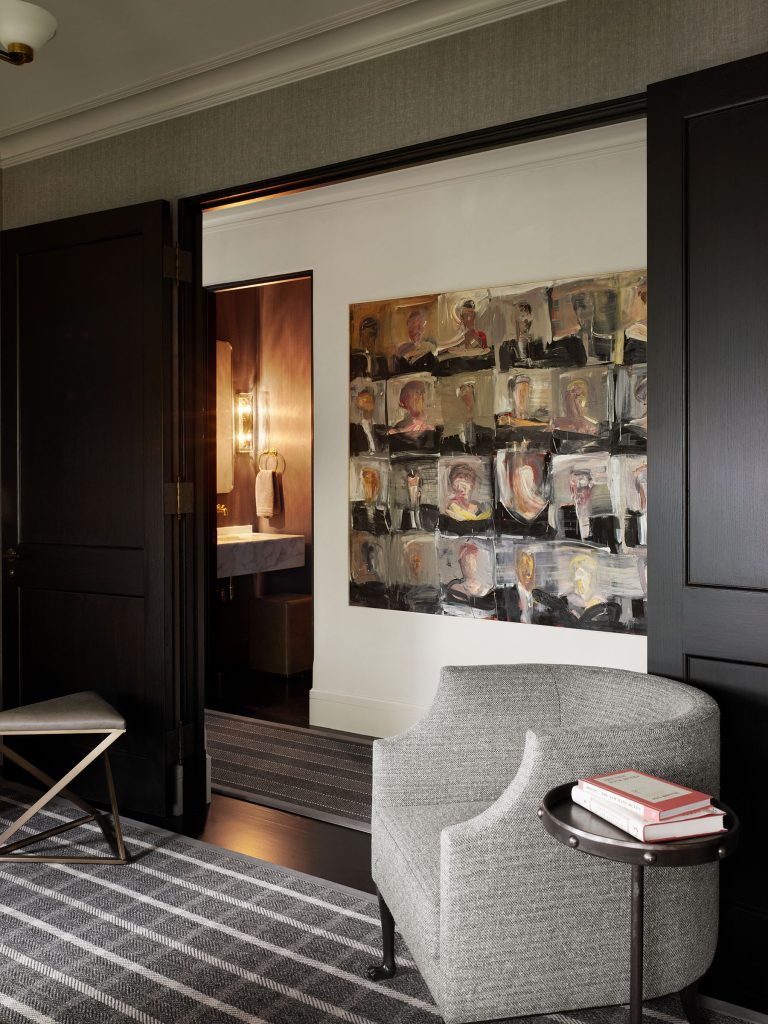 A modern room features dark paneled double doors opening to a bathroom with a lit mirror. On the wall hangs an abstract painting with many faces. In the foreground, there’s a light gray armchair, a round black side table with stacked books, and a striped rug.