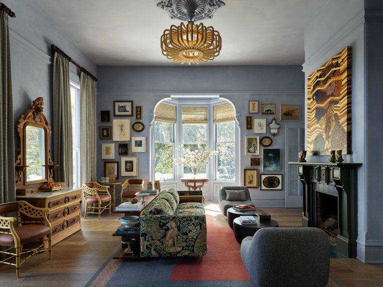 Elegant living room with blue-gray walls, gallery wall of eclectic framed art, floral sofa, two modern gray chairs, golden chairs, vintage wood dresser with mirror, fireplace with art above, large bay window with garden view, wooden floor, and geometric rug.