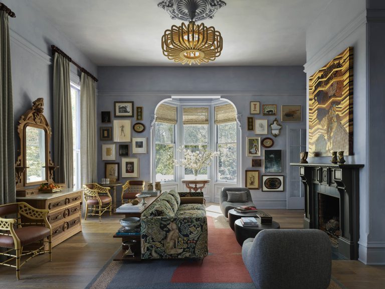 Elegant living room with ornate chandelier, patterned sofa, gray armchairs, and a wooden dresser with a mirror. Artworks fill the pale blue walls. Bay windows with wicker shades let in light. Fireplace with bold abstract art sits across from the main seating area.