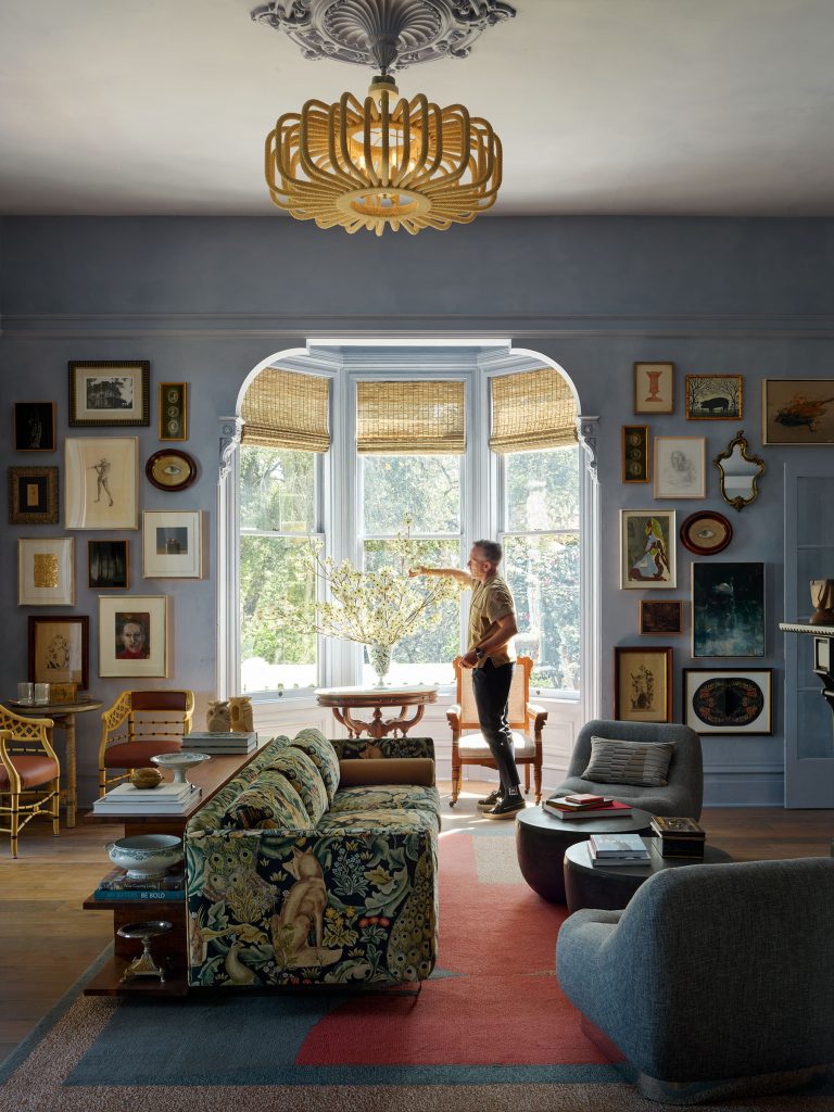 A person arranges flowers in a bay window of an eclectic living room with light blue walls, layered artwork, patterned sofa, gray chairs, a red area rug, modern chandelier, and natural light streaming in through woven blinds.