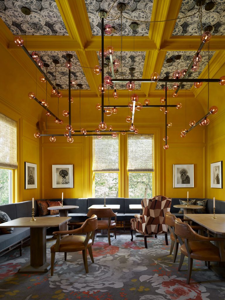 A vibrant room with mustard yellow walls and coffered ceiling, patterned with black-and-white floral wallpaper. Pink orb pendant lights hang above tables and brown chairs. Art prints decorate the walls; a geometric armchair and floral carpet add visual interest.