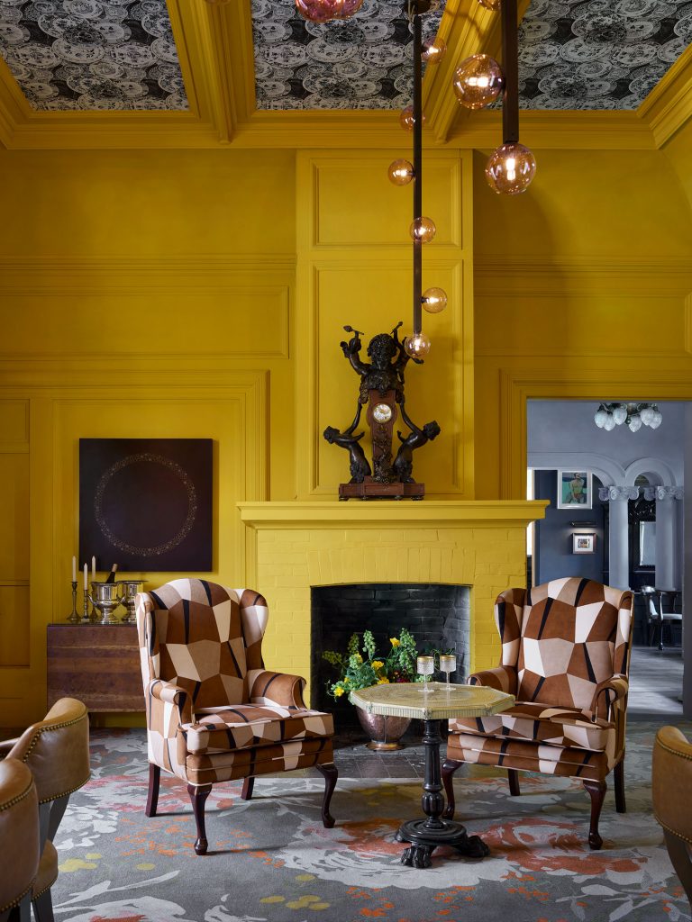 A vibrant room with bright yellow paneled walls and fireplace features two patterned armchairs around a small table. A black statue sits on the mantel. The ceiling is dark with a white pattern, and modern pendant lights hang above. A floral rug covers the floor.