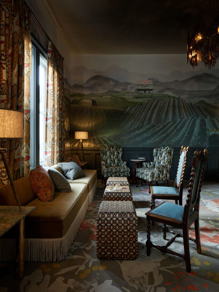 A cozy living room with patterned armchairs, a fringe-trimmed sofa, ottomans, and wooden chairs. Warm light from lamps illuminates the space. The mural on the wall shows green fields, mountains, and a house under a cloudy sky.