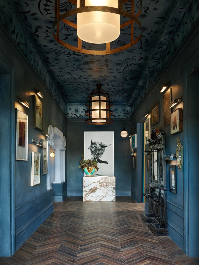 A narrow hallway with dark blue walls and ornate ceiling, decorated with framed art and sculptures. Two large, round pendant lights hang from the ceiling. At the end, a marble-topped console and a dynamic black horse artwork are visible. The floor is herringbone wood.