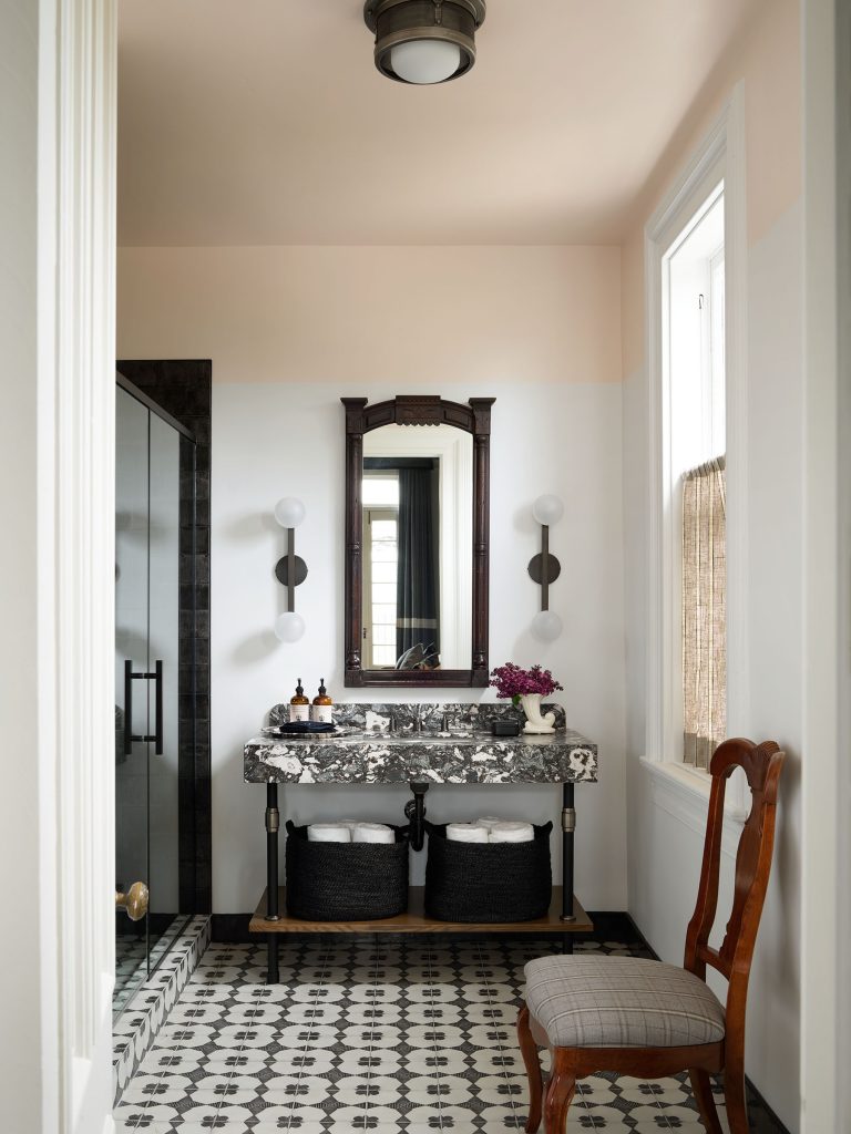 A bathroom with patterned black-and-white tile floor, marble-topped vanity with two black baskets, two bottles, and a vase of flowers. Above the vanity is a mirror flanked by two wall lights. A shower with glass door is on the left, and a wooden chair sits on the right.