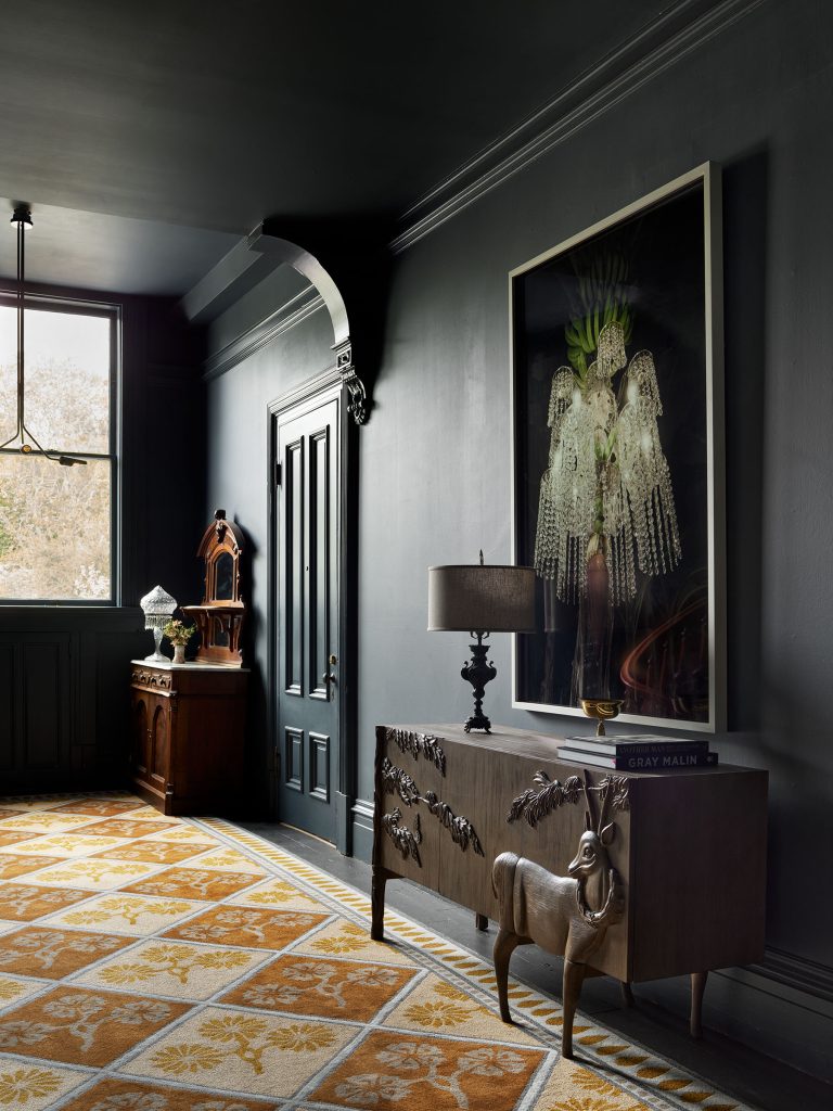 A moody, dark-walled room features a carved sideboard with a sculpted animal, a lamp, a large portrait of a figure in ornate costume, a book, a vintage clock, and golden patterned carpet, with light streaming through a window.