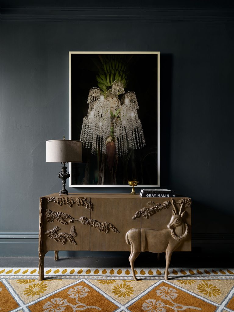 A dark wall features a large framed photo of a crystal chandelier above a carved wooden console with branch and deer motifs. On the console are a white lamp, a glass of wine, and a book. A patterned rug with yellow and white accents is on the floor.