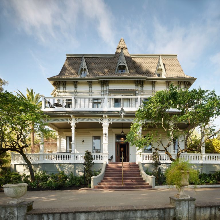 A large, ornate Victorian-style house with a steep gabled roof, decorative trim, and white wood siding. It features a broad porch with balustrades, upper balcony, symmetrical windows, and a central staircase leading to the front door, surrounded by green trees and shrubs.