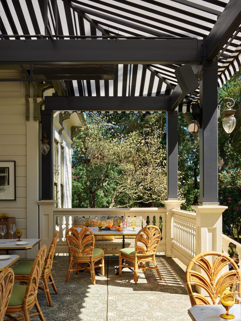A sunny patio with striped black-and-white awning, rattan chairs with green cushions, white tables, and a plant-filled view. Sunlight filters through the trees and awning, casting shadows on the patterned tile floor. Elegant, outdoor dining setting.