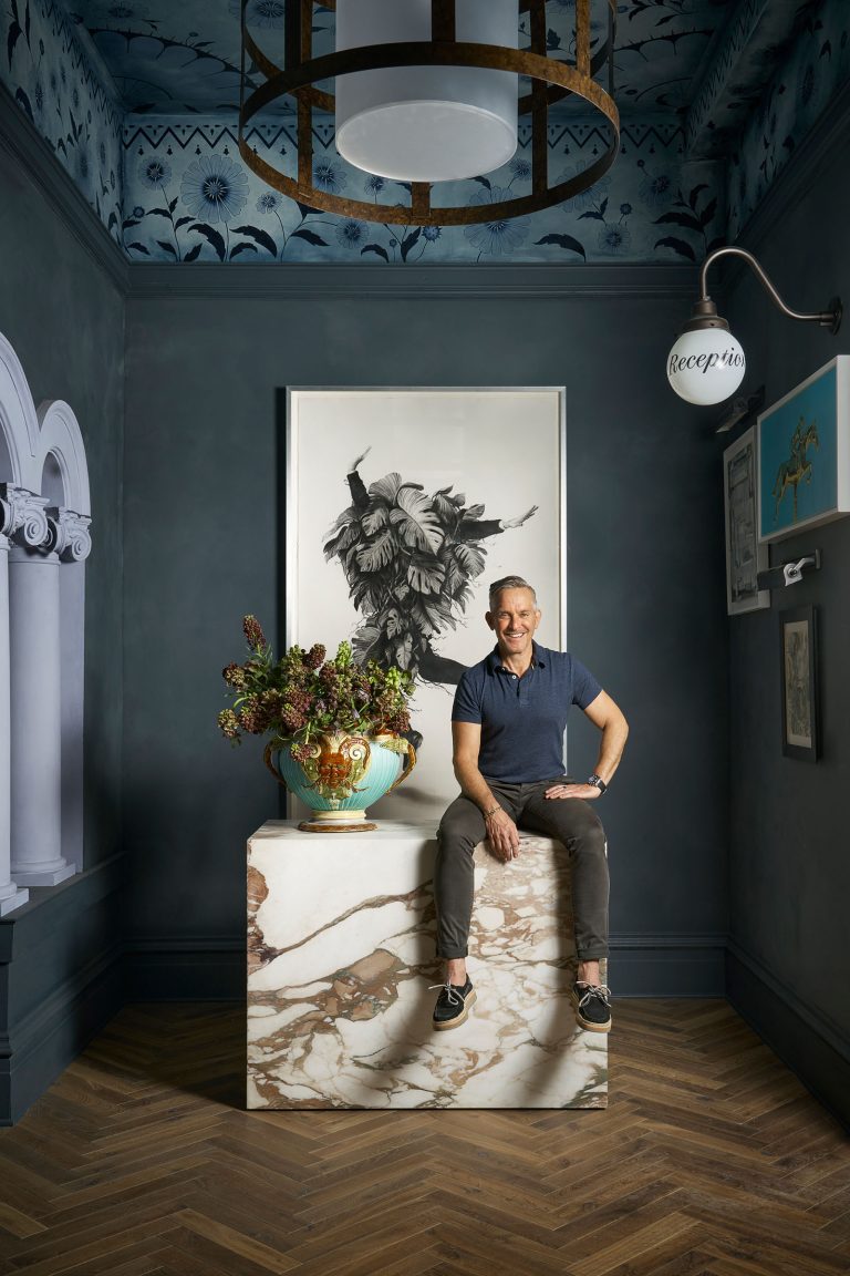 A smiling man in a navy shirt and gray pants sits on a large marble pedestal beside a floral arrangement in a decorative vase, beneath black-and-white art. The ornate room features dark blue walls, patterned ceiling, wood floor, and a “receptio” wall sign.