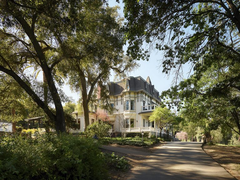 A large, ornate Victorian-style house with a wraparound porch sits surrounded by tall, leafy trees and lush greenery. Sunlight filters through the branches, and a paved path curves toward the house, creating a serene, inviting scene.