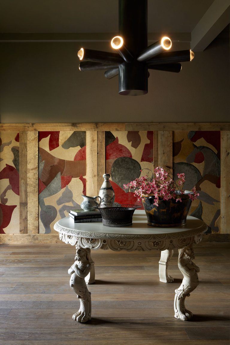 An ornate white table with carved animal legs stands on a wooden floor. On the table are ceramic dishes and a dark vase holding pink flowers. Behind is a colorful abstract mural in red, yellow, and gray tones, and a modern black chandelier hangs above.