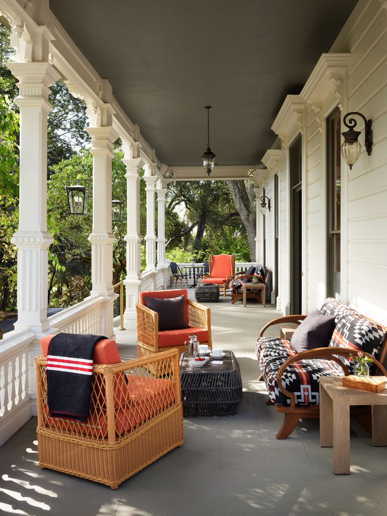 A Victorian-style porch with white ornate columns features wicker chairs with orange cushions, a patterned black-and-white sofa, a small table, throw blankets, and pillows. Hanging lantern lights and greenery are visible in the background.