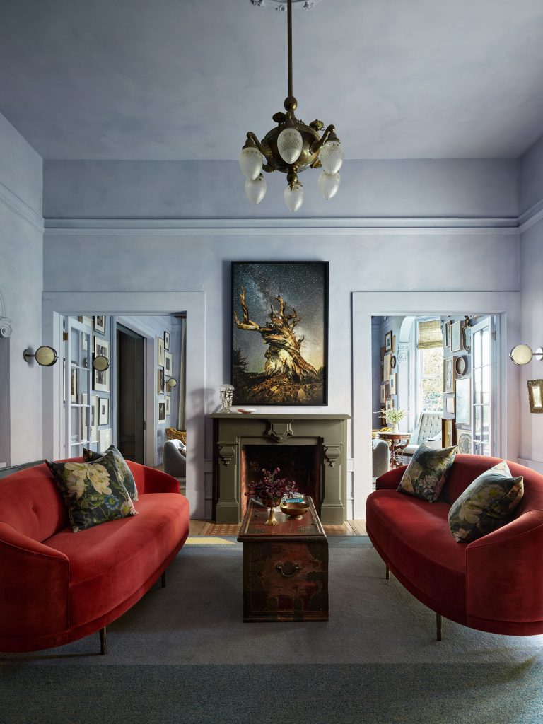 A stylish living room with two red velvet sofas facing each other, each with floral pillows. A vintage trunk serves as a coffee table. A fireplace centers the back wall, topped with dramatic artwork of a tree, and a classic chandelier hangs from the ceiling.