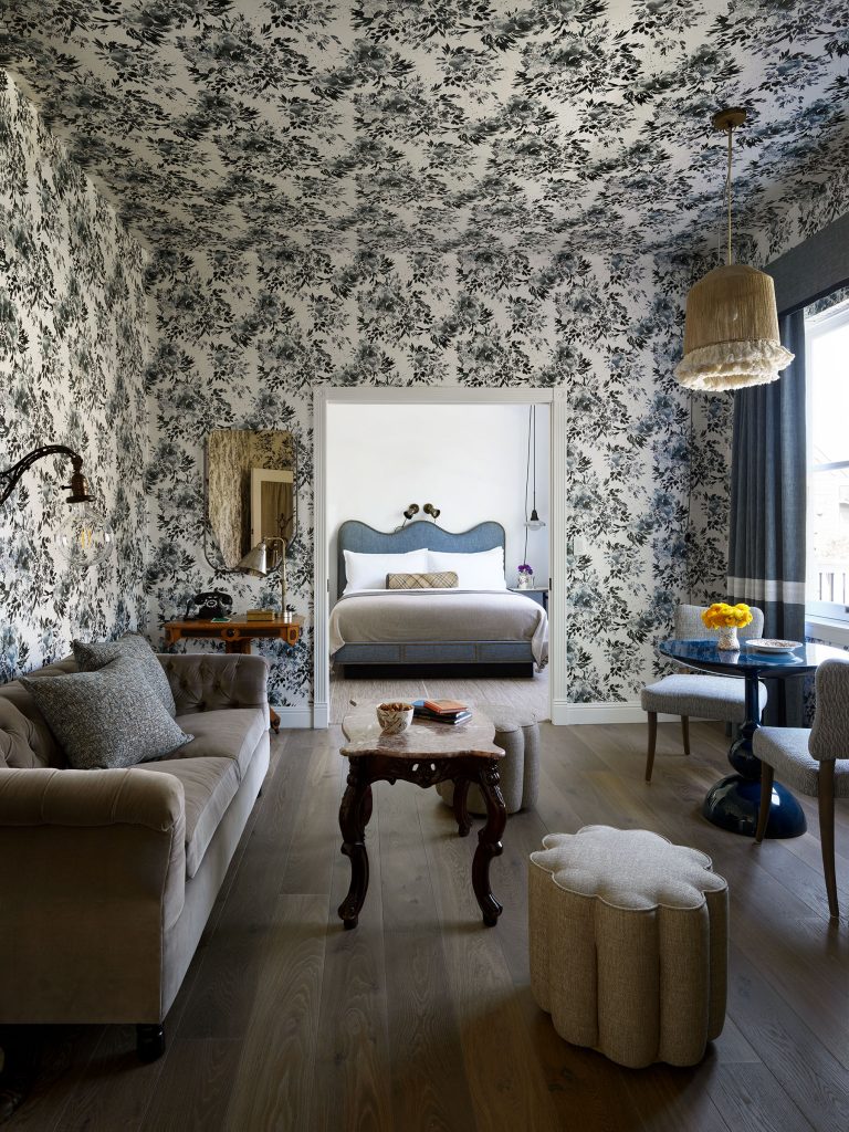 A stylish room with black and white floral wallpaper covering the walls and ceiling, a bed with a blue scalloped headboard, a beige tufted sofa, ornate wood coffee table, plush ottoman, round dining table, and large sunny window with a hanging fringed lamp.