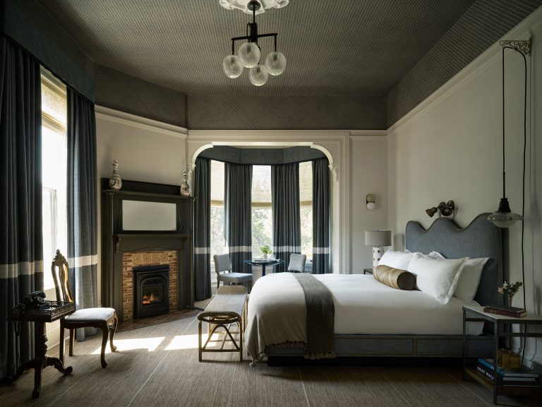 A stylish bedroom with high ceilings, a gray upholstered bed, white bedding, and a brown throw. There is a brick fireplace, bay windows with dark curtains, a small round table with two chairs, vintage décor, and soft natural light filling the room.