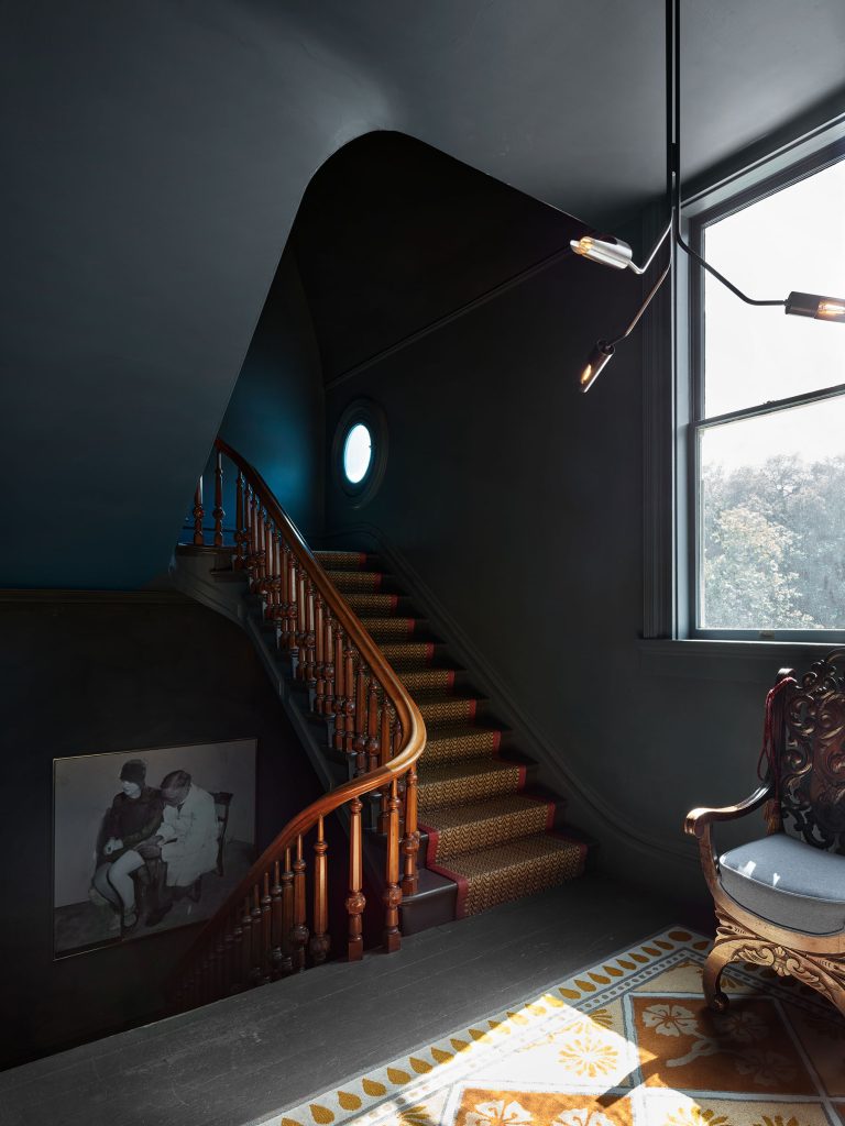 A dark-walled staircase with a wooden banister curves upward, featuring patterned carpet. A round window and a large rectangular window let in natural light. A vintage armchair and ornate rug sit nearby; a black-and-white photo hangs on the wall by the stairs.
