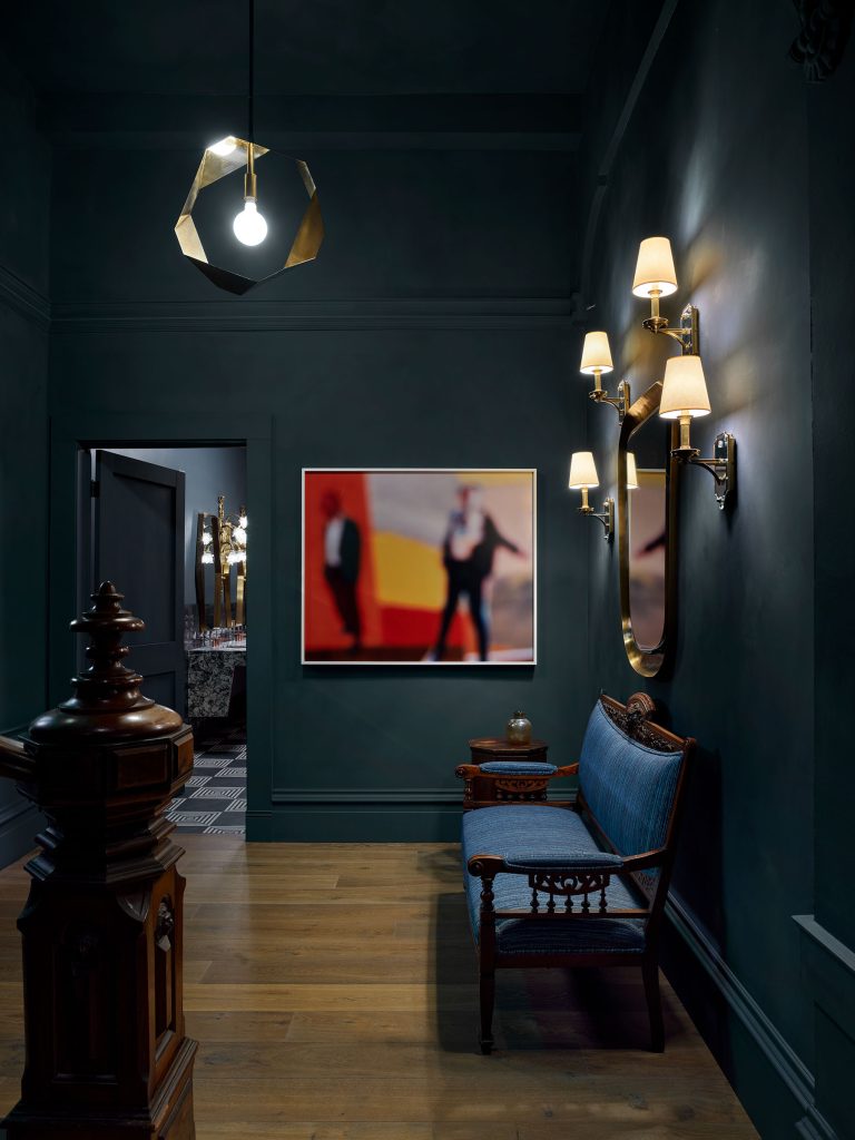 A dim hallway with dark teal walls, wooden floor, and ornate bench. Modern pendant light and classic wall sconces with shades illuminate the space. A large, blurred red and yellow abstract photo and a gold-framed mirror decorate the wall.