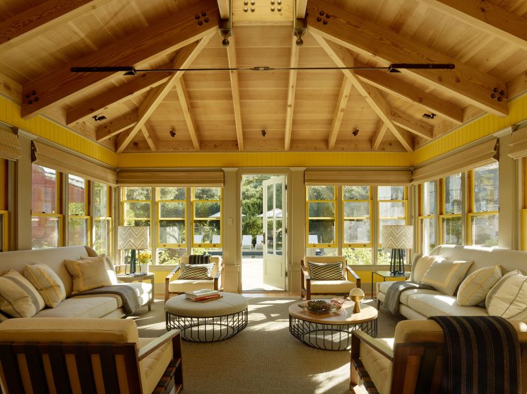 A bright sunroom with a vaulted wood ceiling, exposed beams, and yellow walls. Large windows line the sides, letting in natural light. Neutral sofas, round ottomans, and a coffee table are arranged around a rug. Double doors open to a sunlit patio.