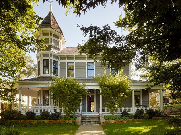 A two-story Victorian house with a prominent turret, wraparound porch, gray siding, and white trim. Mature trees and landscaping frame the front, and sunlight filters through the leaves, casting dappled shadows on the house and walkway.
