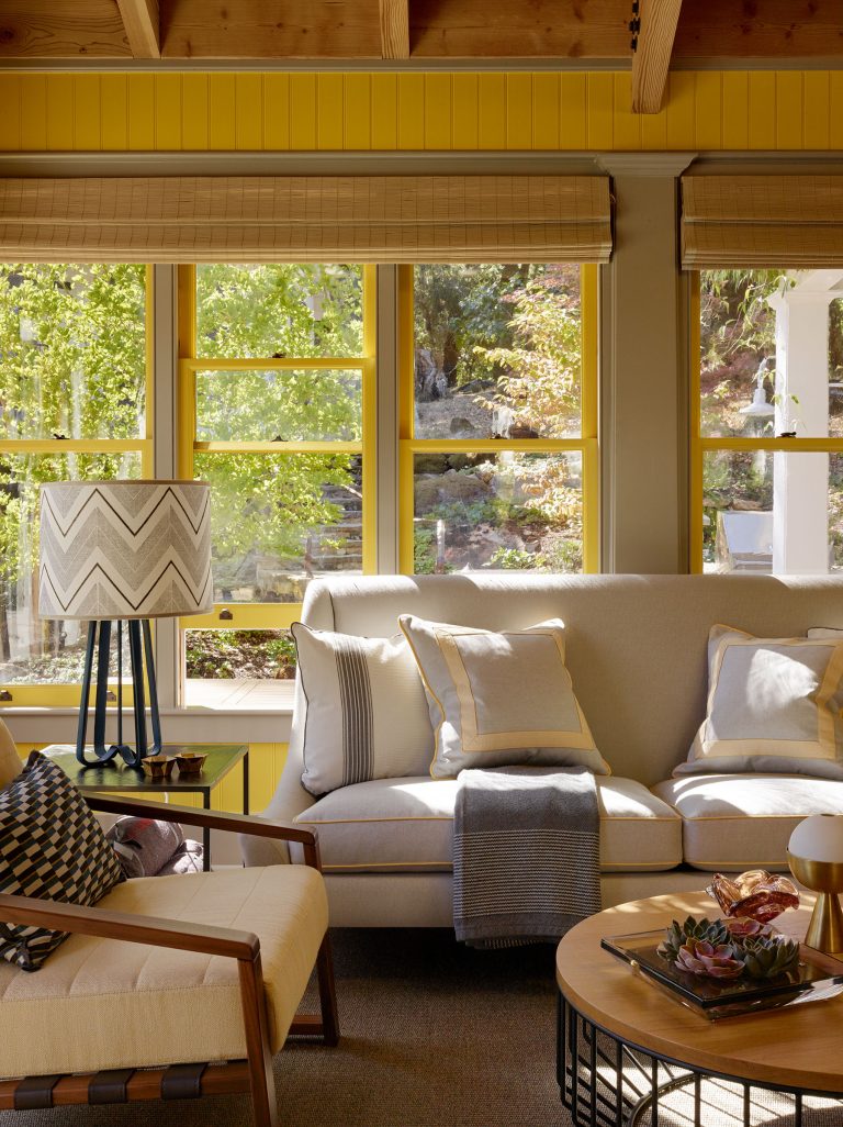 A cozy living room featuring a cream sofa with pillows, a gray throw, and a wooden armchair. A chevron-patterned lamp sits on a yellow side table. Sunlight streams through large windows with yellow trim, illuminating a coffee table with a succulent plant.