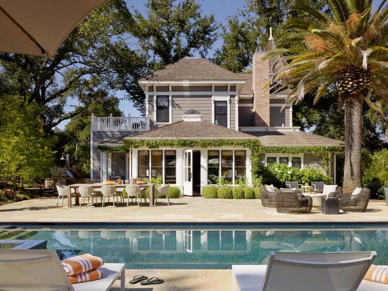 A gray two-story house with white trim sits behind a patio with a dining table and lounge chairs, surrounded by lush greenery and a tall palm tree. In the foreground, a swimming pool reflects the house and blue sky. Towels, sandals, and shaded loungers are poolside.