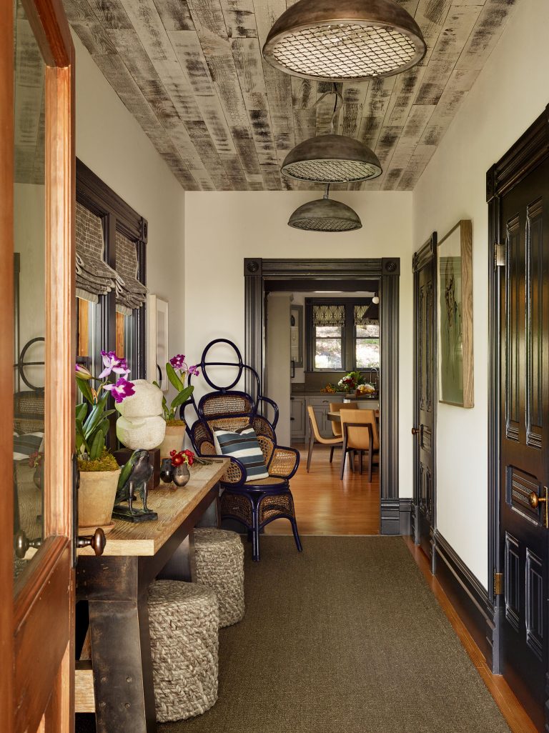 A hallway with rustic wood ceiling, dark trim, and light walls features a wooden console table, potted orchids, decorative stools, a large mirror, and patterned chairs. Pendant lights hang above, and a dining area is visible at the end of the hallway.