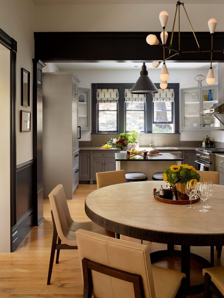 A modern kitchen with gray cabinets, stainless appliances, and a round dining table with beige chairs. Sunflowers and wine glasses sit on the table. A large window brings in light, and pendant lights hang from the ceiling, illuminating the space warmly.
