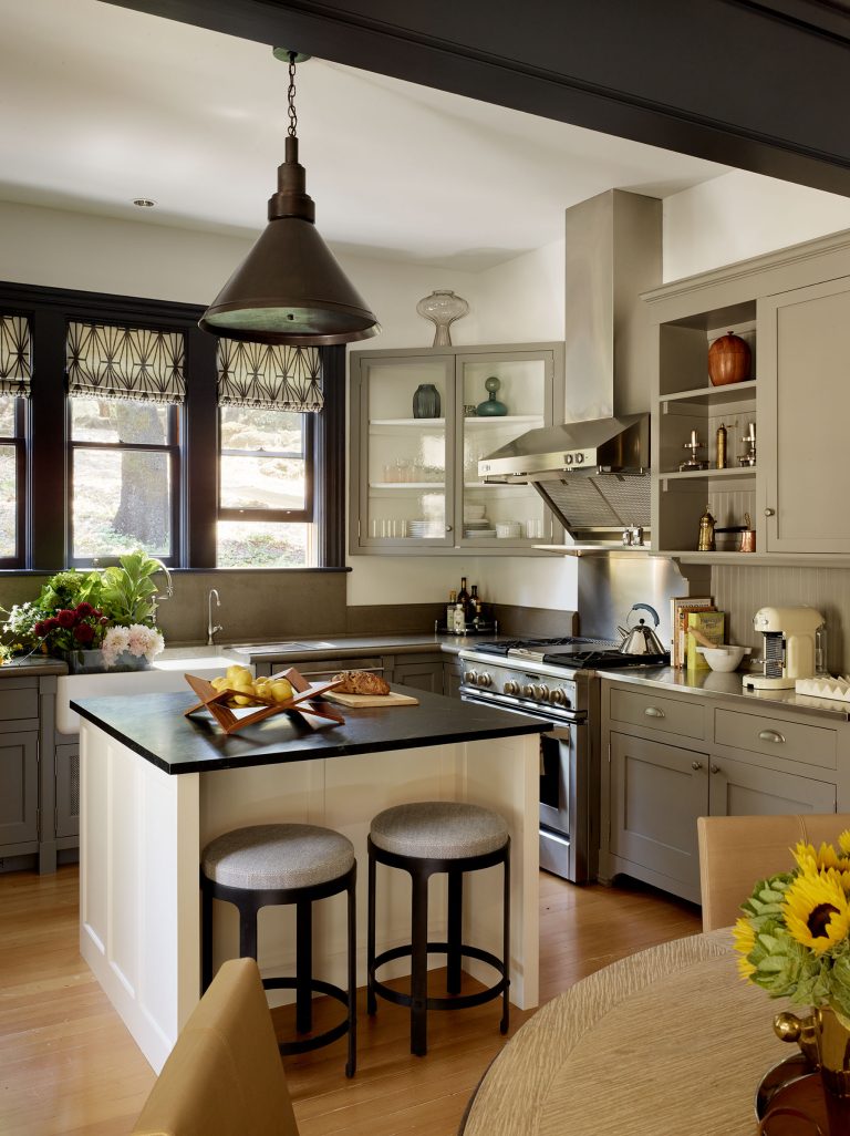 Modern kitchen with gray cabinets, open shelves, and stainless steel appliances. An island with two cushioned stools sits in the center, with bread and cutting boards on top. Large windows with patterned valances let in light; flowers and fresh produce decorate the space.