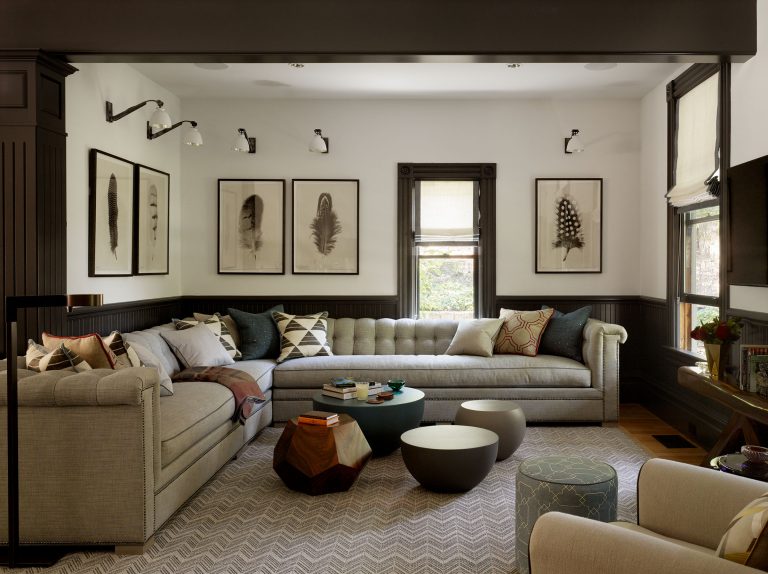 A cozy living room with two beige tufted sofas, round and hexagonal coffee tables, patterned pillows, feather art prints on the wall, chevron rug, dark wainscoting, modern lighting, and windows with neutral roman shades letting in natural light.