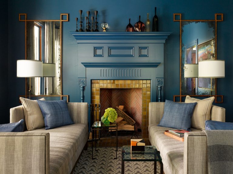 A stylish living room with deep blue walls features two beige sofas facing each other, a blue fireplace with decorative vases, mirrors on either side, wall-mounted lamps, patterned rug, glass tables, and a fireplace filled with stacked logs and green flowers.