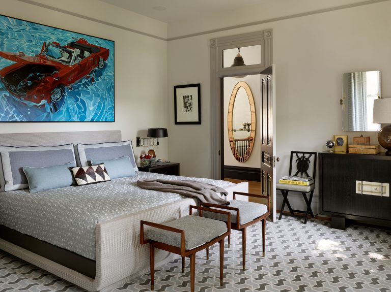 A modern bedroom with a patterned gray rug, upholstered bed with gray bedding, two wooden stools, and abstract art of a red car above the bed. A sleek black dresser, decorative objects, and a doorway with an oval mirror are also visible.