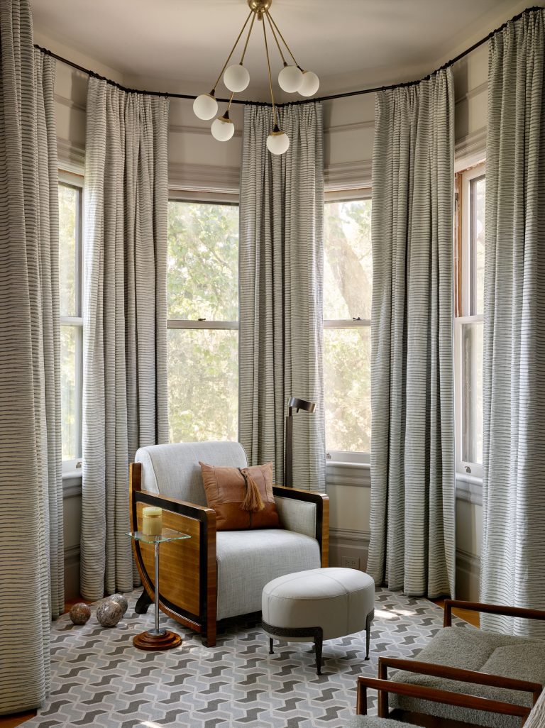 A cozy corner features a modern armchair with wooden arms, a light ottoman, and brown cushions. Floor-to-ceiling gray curtains frame bay windows with leafy views. A geometric rug and a brass chandelier with globe lights complete the serene setting.