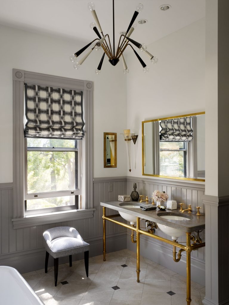 Elegant bathroom with gray wainscoting, gold fixtures, a double sink with exposed gold plumbing, a large gold-framed mirror, a small upholstered chair near a window with a patterned blind, and a modern black-and-gold chandelier overhead.