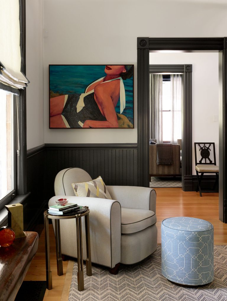 A cozy reading nook with a light gray armchair, a small round side table, and a blue patterned ottoman. The walls are white with black trim and wainscoting. A large painting of a reclining woman hangs above, and a window lets in natural light.