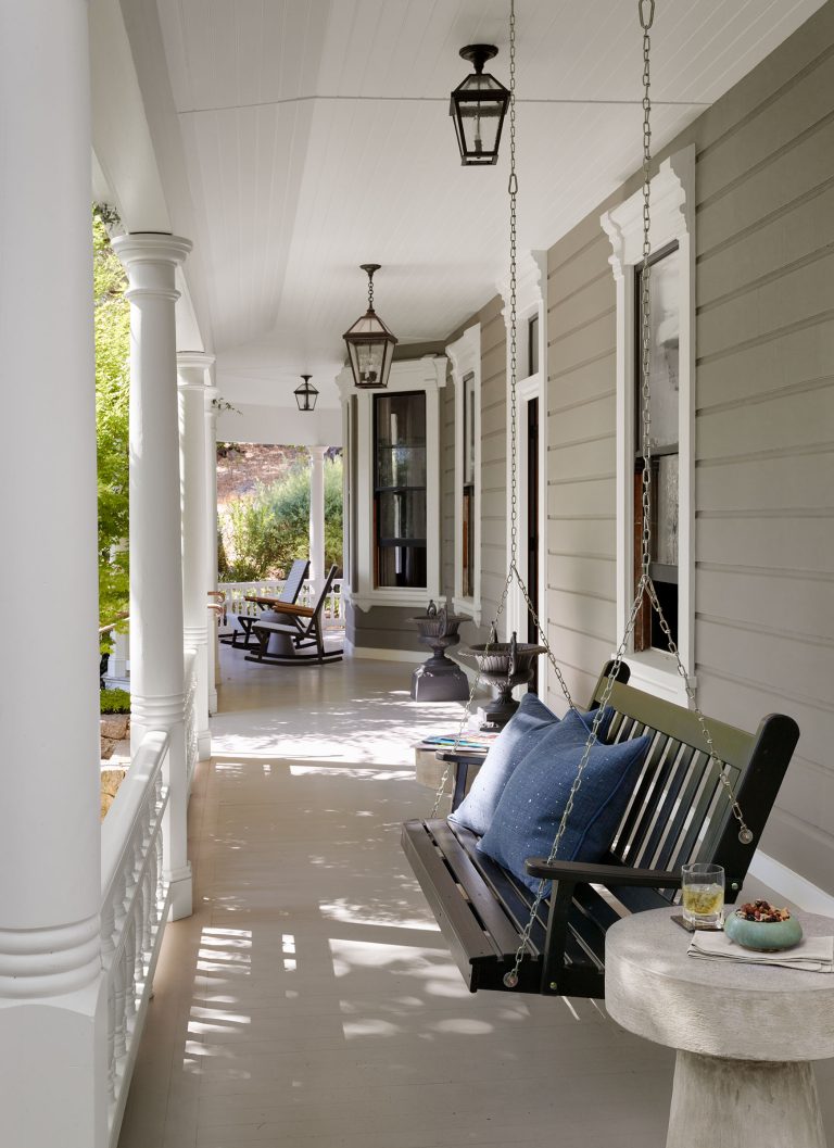 A shaded porch with white columns, gray siding, and a hanging black bench swing with blue pillows. A small round table holds a drink and snack. Lantern lights hang from the ceiling; rocking chairs and greenery are visible in the background.