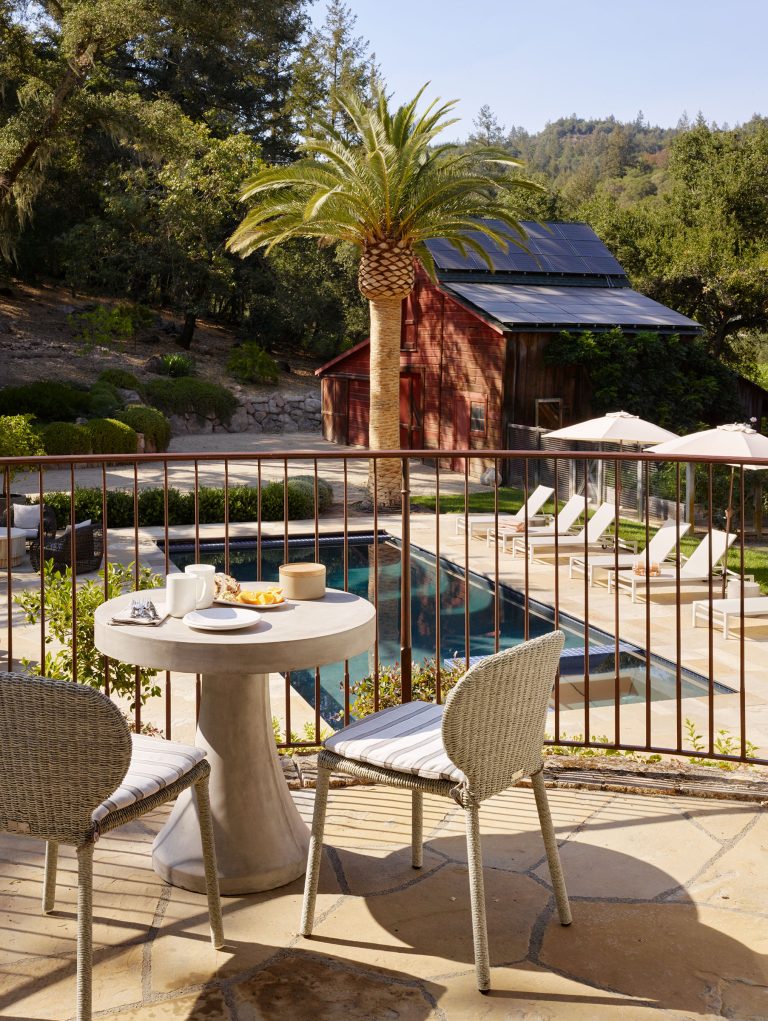 A small round table with two chairs sits on a patio overlooking a pool, sun loungers, a red barn, and a palm tree. Hills with lush greenery and trees are visible in the background under a clear sky. Breakfast items are on the table.