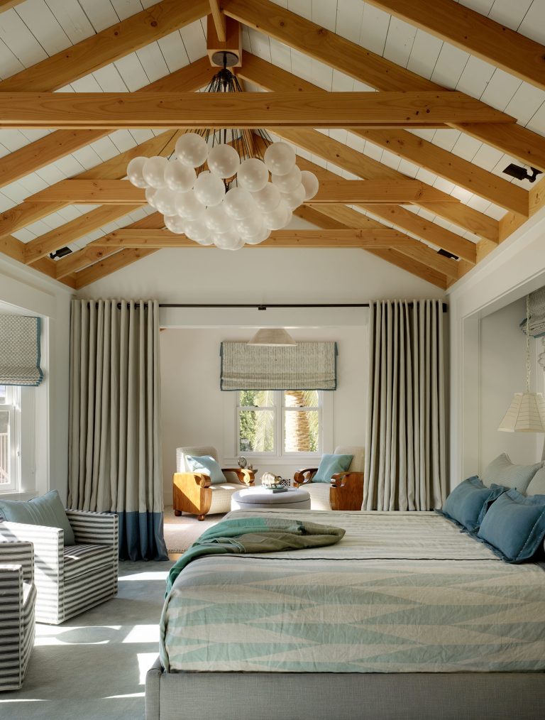 A cozy bedroom with a wood-beamed vaulted ceiling, modern bubble chandelier, and large windows. The space features a gray bed with striped bedding, striped armchairs, and a sitting area with armchairs and blue cushions, creating a serene, airy atmosphere.