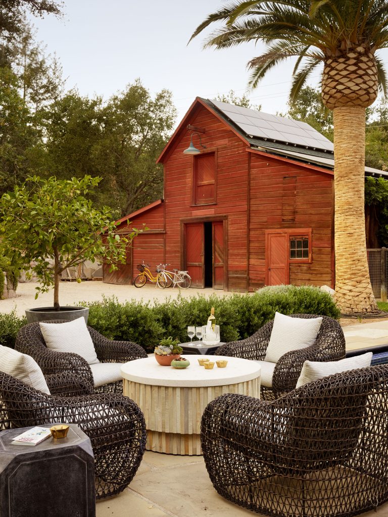 Four black wicker armchairs with white cushions surround a round table outdoors. On the table are drinks and a plant. Behind is a red wooden barn, two yellow bikes, a lemon tree, a palm, and greenery, creating a relaxed, rustic setting.