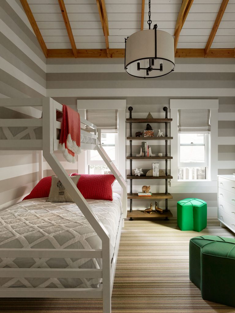 A cozy bedroom with a white bunk bed, gray and white striped walls, red pillows, and a red blanket. There’s a striped rug, green ottomans, a dark bookshelf with decor, large windows, white drawers, and a modern drum pendant light. Exposed wooden beams on the ceiling.