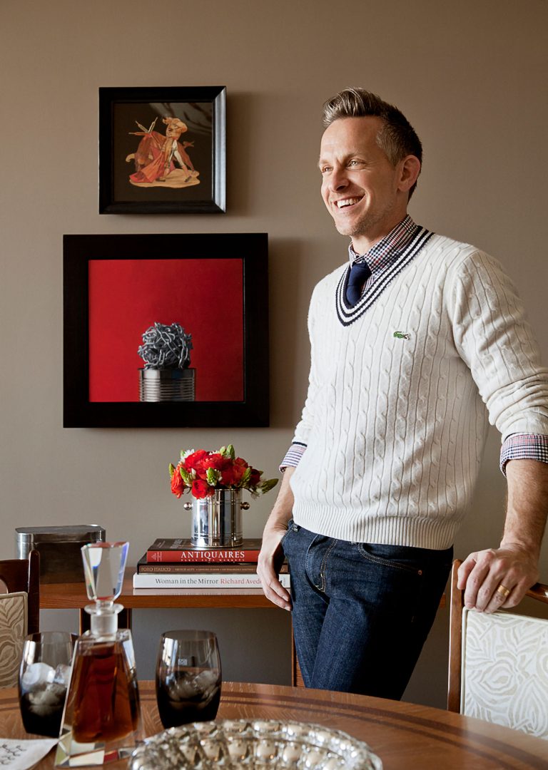 A smiling man in a white cable-knit sweater and dark jeans stands by a dining table with books, flowers, and drinks. Behind him, a beige wall displays two art pieces: a figure on top and a can with a dark object against a red background below.