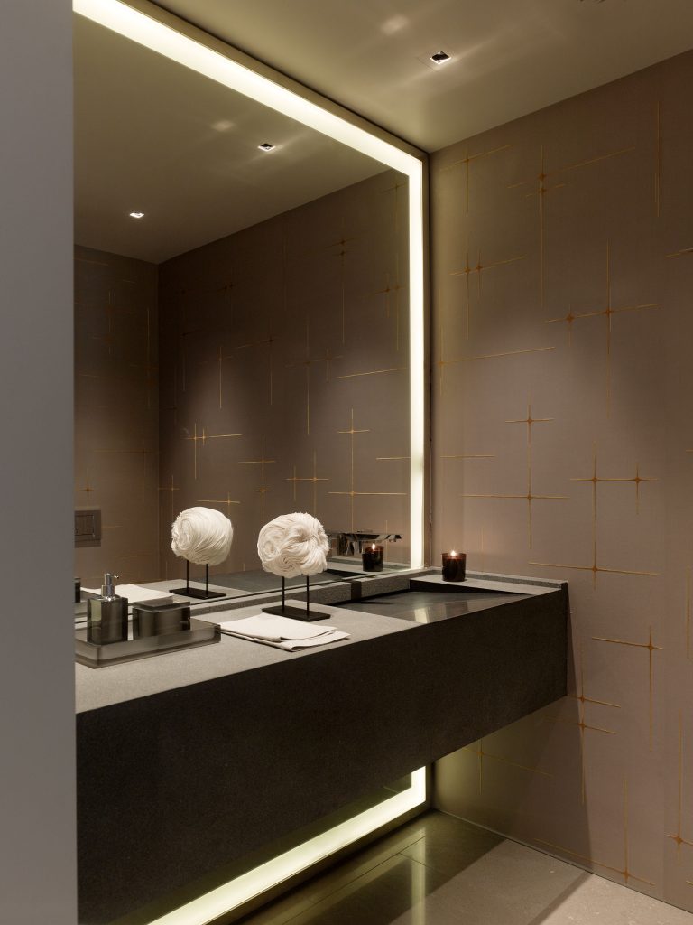 A modern bathroom with a sleek gray floating vanity, double sinks, and a large backlit mirror. The walls are decorated with a matte finish and gold line patterns. Minimalist accessories and sculptural white decor pieces sit on the countertop.