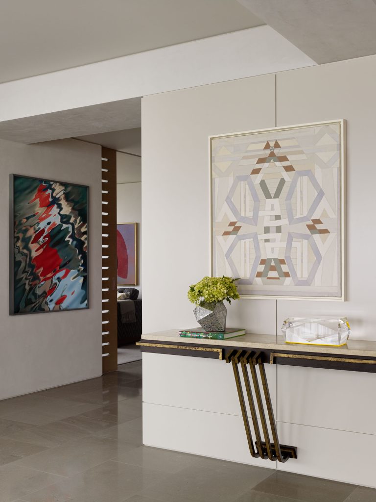 Modern foyer with glossy gray floor tiles, a floating black and gold console table holding a green floral arrangement and a crystal tray. Abstract geometric artwork hangs above the table; another colorful abstract painting is on the wall in the hallway to the left.