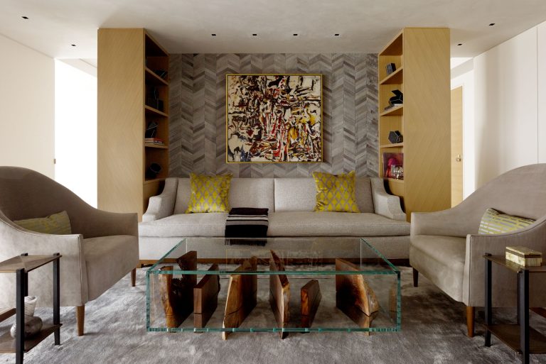 Contemporary living room with gray sofa, yellow-patterned pillows, two beige armchairs, a glass coffee table with wooden accents, built-in shelving, abstract art on a herringbone-patterned wall, neutral tones, and soft carpet flooring.