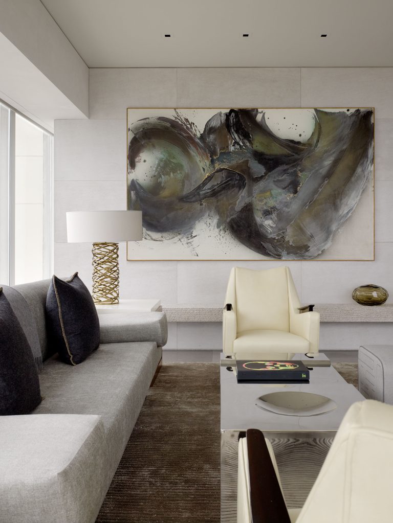 A modern living room with a large abstract painting on a light wall, a gray sofa with dark pillows, two cream chairs, a white coffee table with books, a gold lamp, and a glass bowl. The space is bright with natural light from a window.